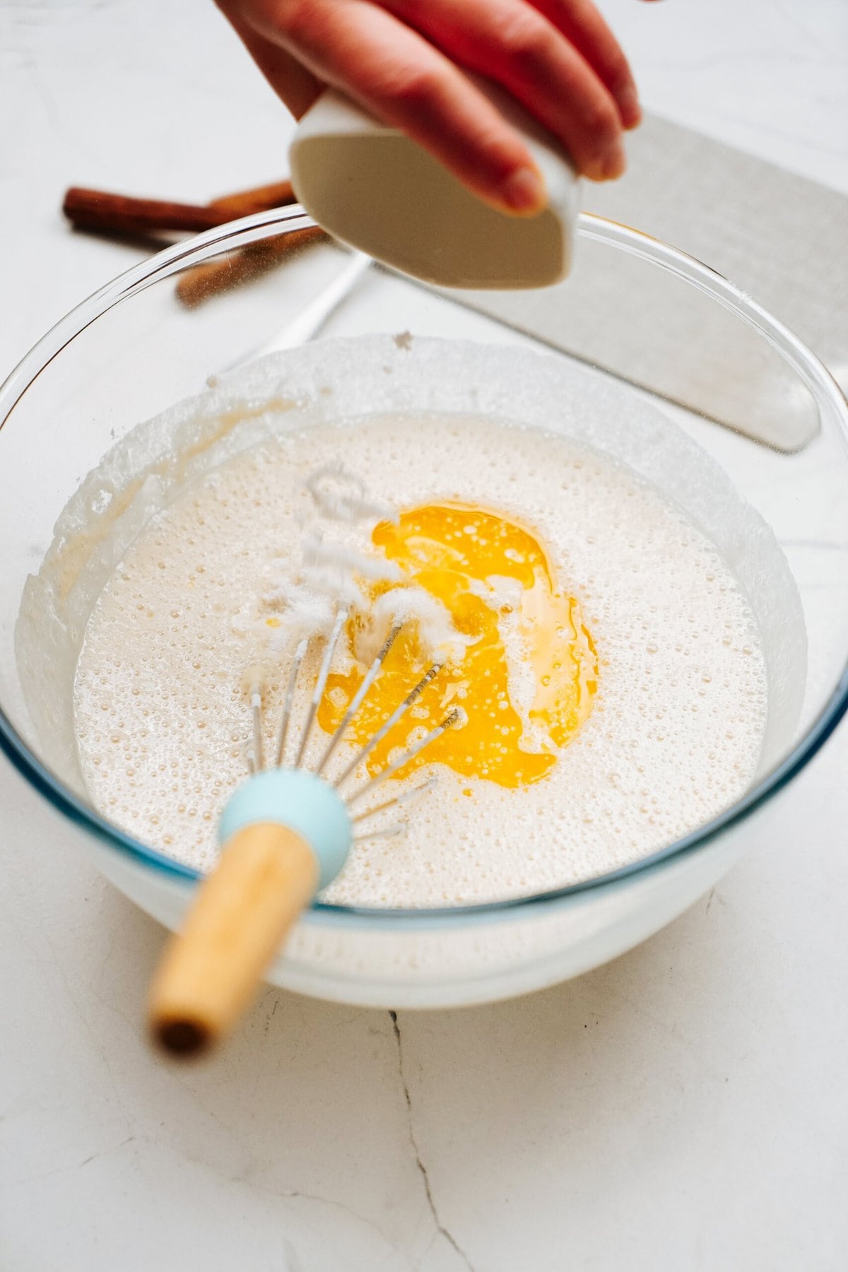 A hand is adding sugar from a small ceramic container into a glass bowl containing whisked eggs and flour, perfect for making cinnamon rolls. There is a whisk inside the bowl while a cinnamon stick and a grater lay on the counter.