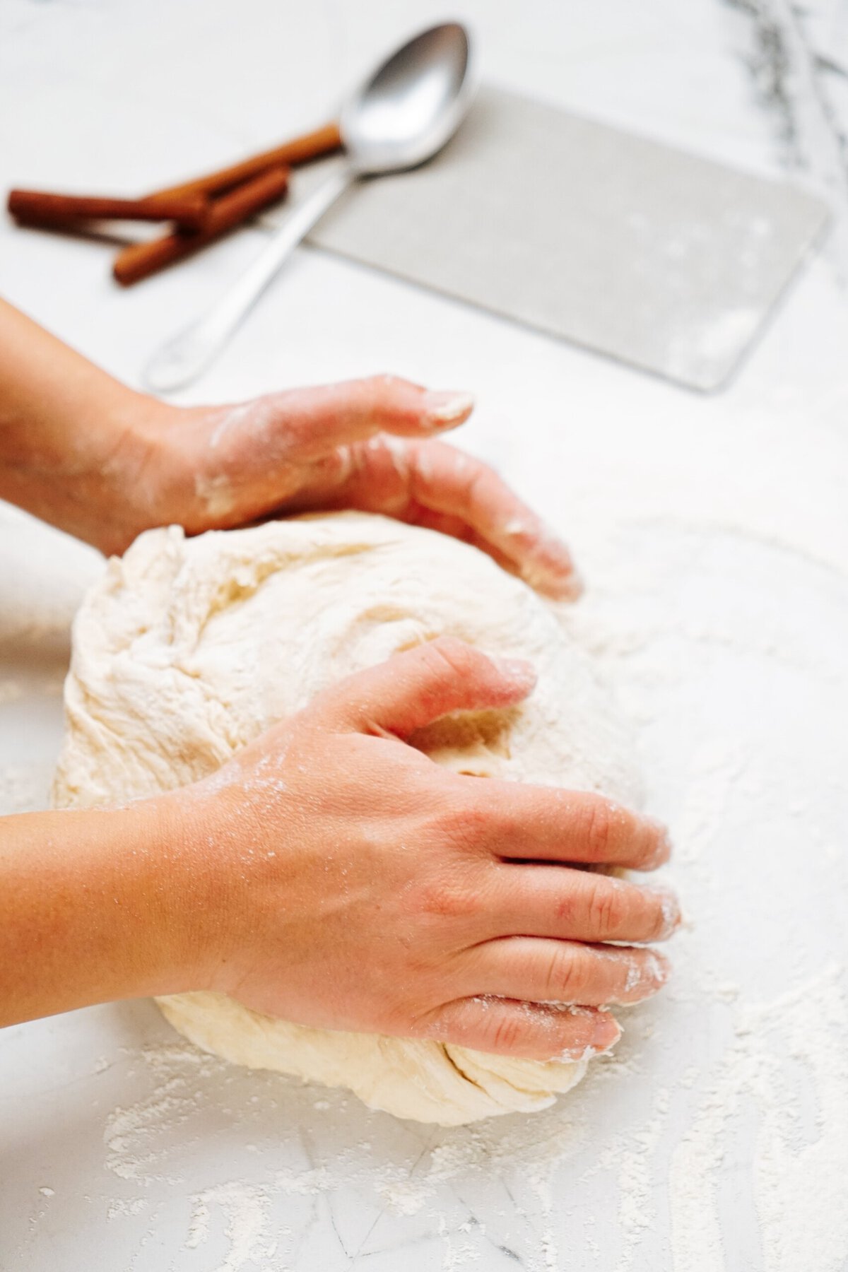 Hands kneading a ball of dough on a floured surface, preparing for delicious cinnamon rolls, with a spoon, cutting board, and cinnamon sticks in the background.
