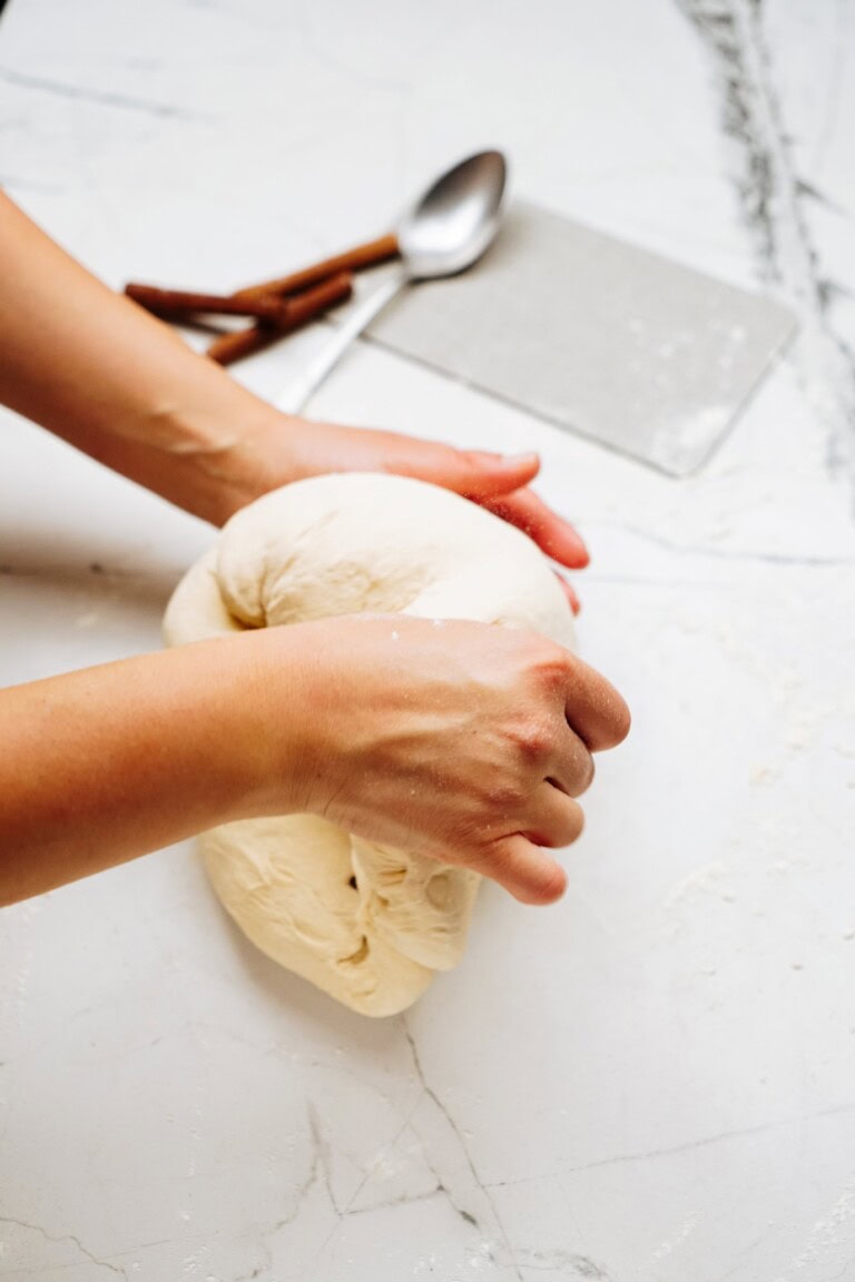 Two hands knead a ball of dough on a marble countertop, preparing the base for delectable cinnamon rolls. A spoon and two sticks of cinnamon are visible in the background, hinting at the aromatic treat to come.