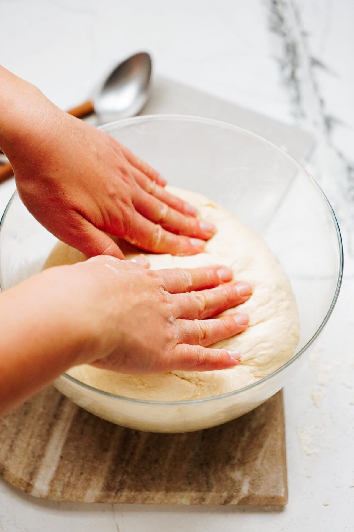 Hands pressing down on dough in a glass bowl, with a spoon on the side; photo taken on a marble surface. The preparation of cinnamon rolls is well underway.