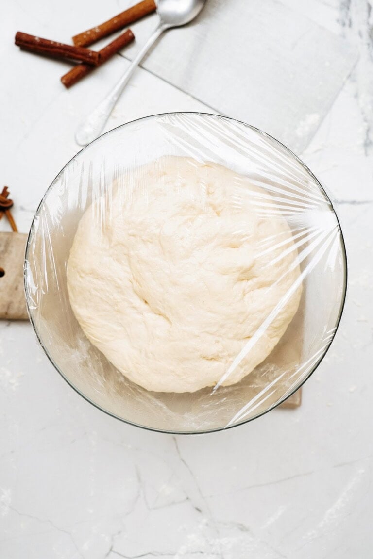 A glass bowl filled with dough, soon to become delicious cinnamon rolls, rests on a marble surface covered in plastic wrap. A spoon and cinnamon sticks are in the background.