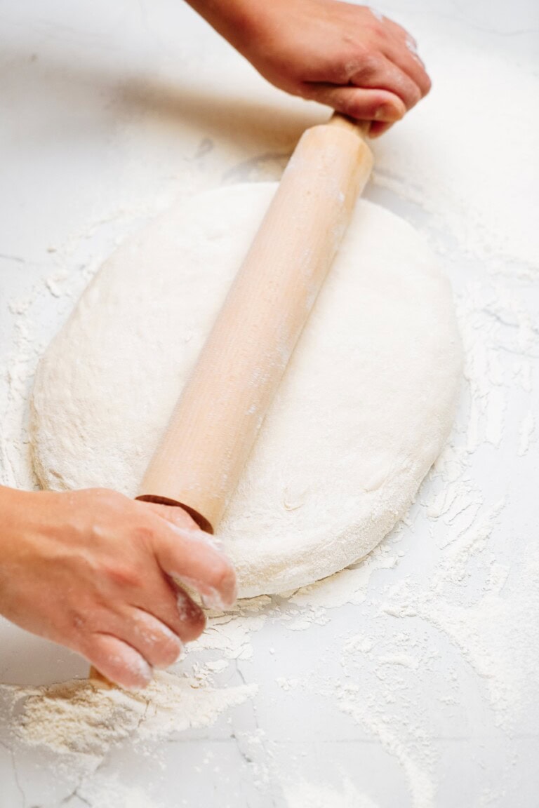 Hands rolling out dough with a wooden rolling pin on a floured surface, preparing the perfect base for mouthwatering cinnamon rolls.