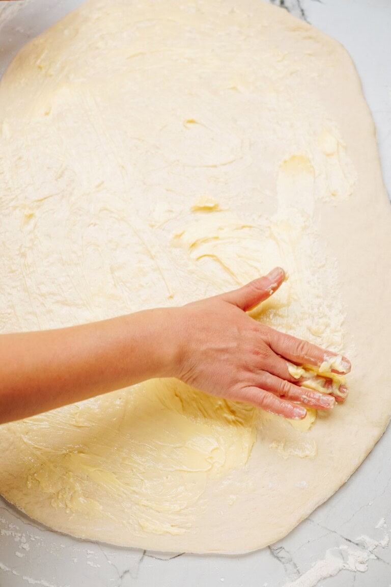 Close-up of a person's hand spreading a layer of butter onto a large, flat dough on a floured surface, preparing the perfect base for delicious cinnamon rolls.