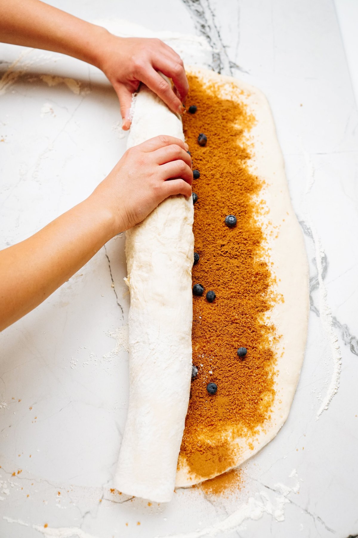 Person rolling dough with cinnamon filling and blueberries on a white marble surface, preparing delicious cinnamon rolls.