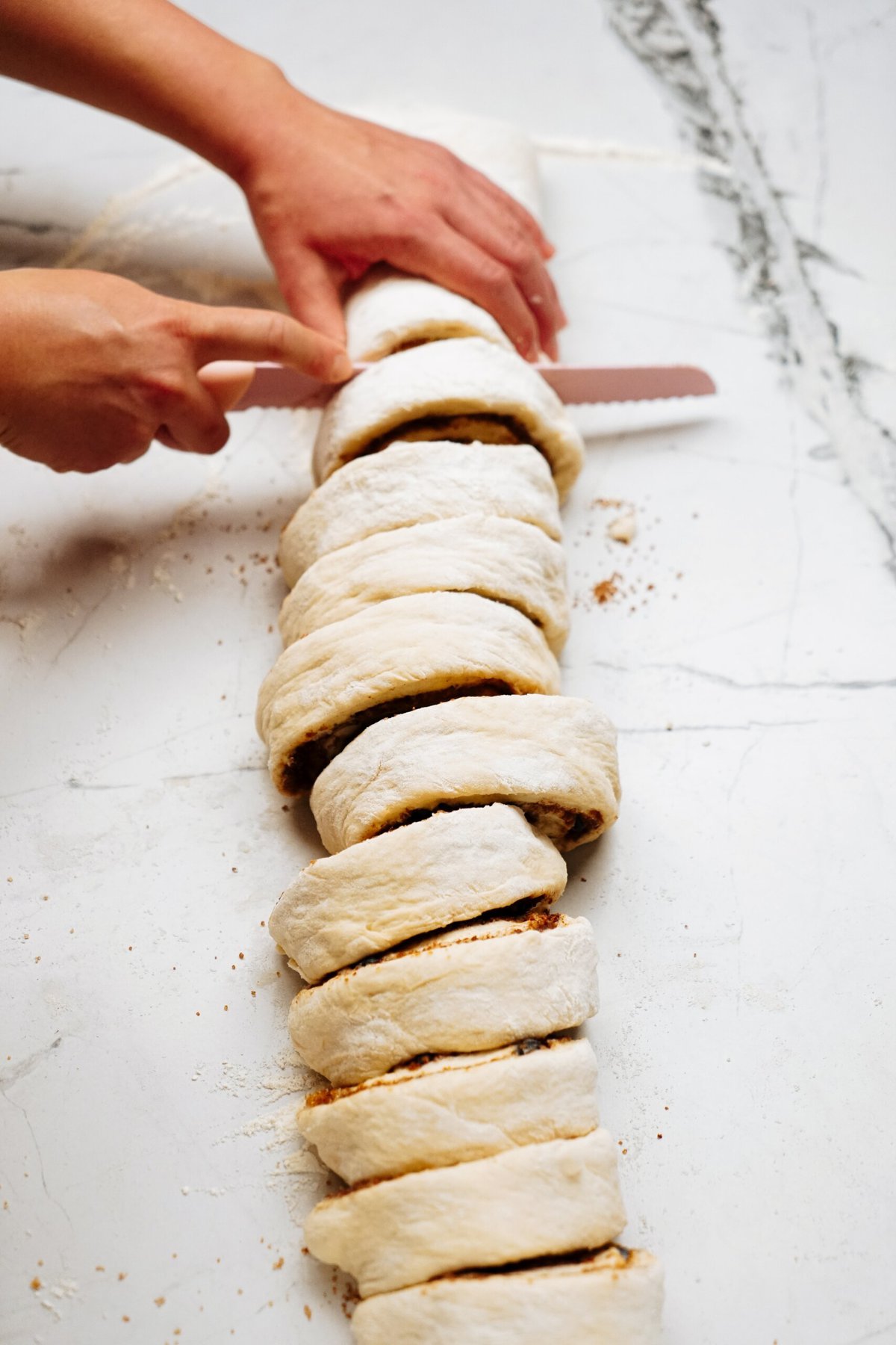 Hands cutting rolled dough with a serrated knife into even portions on a white marble surface, preparing delicious cinnamon rolls.