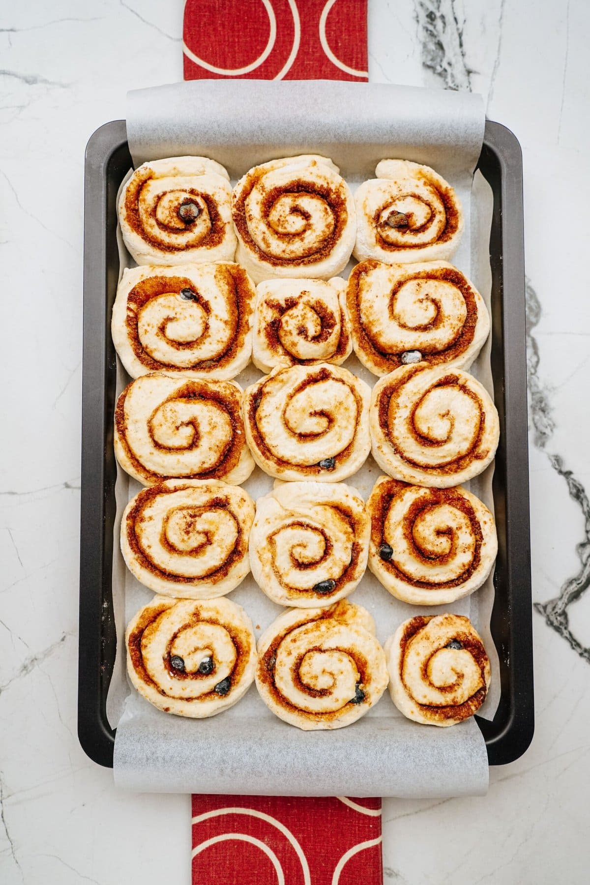A baking tray featuring evenly spaced, uncooked cinnamon rolls on parchment paper, arranged in a 4x5 grid pattern, rests on a marble countertop with a red and white patterned cloth underneath.