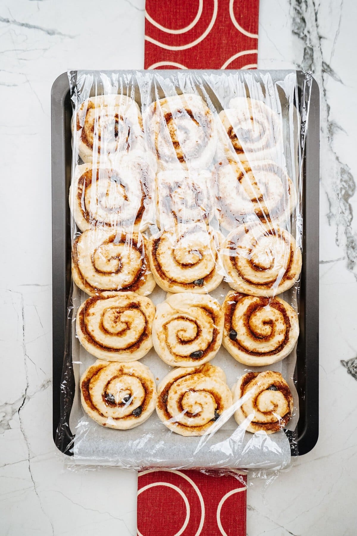 A baking sheet with a dozen uncooked cinnamon rolls sits on a marble countertop, each roll carefully covered in plastic wrap.