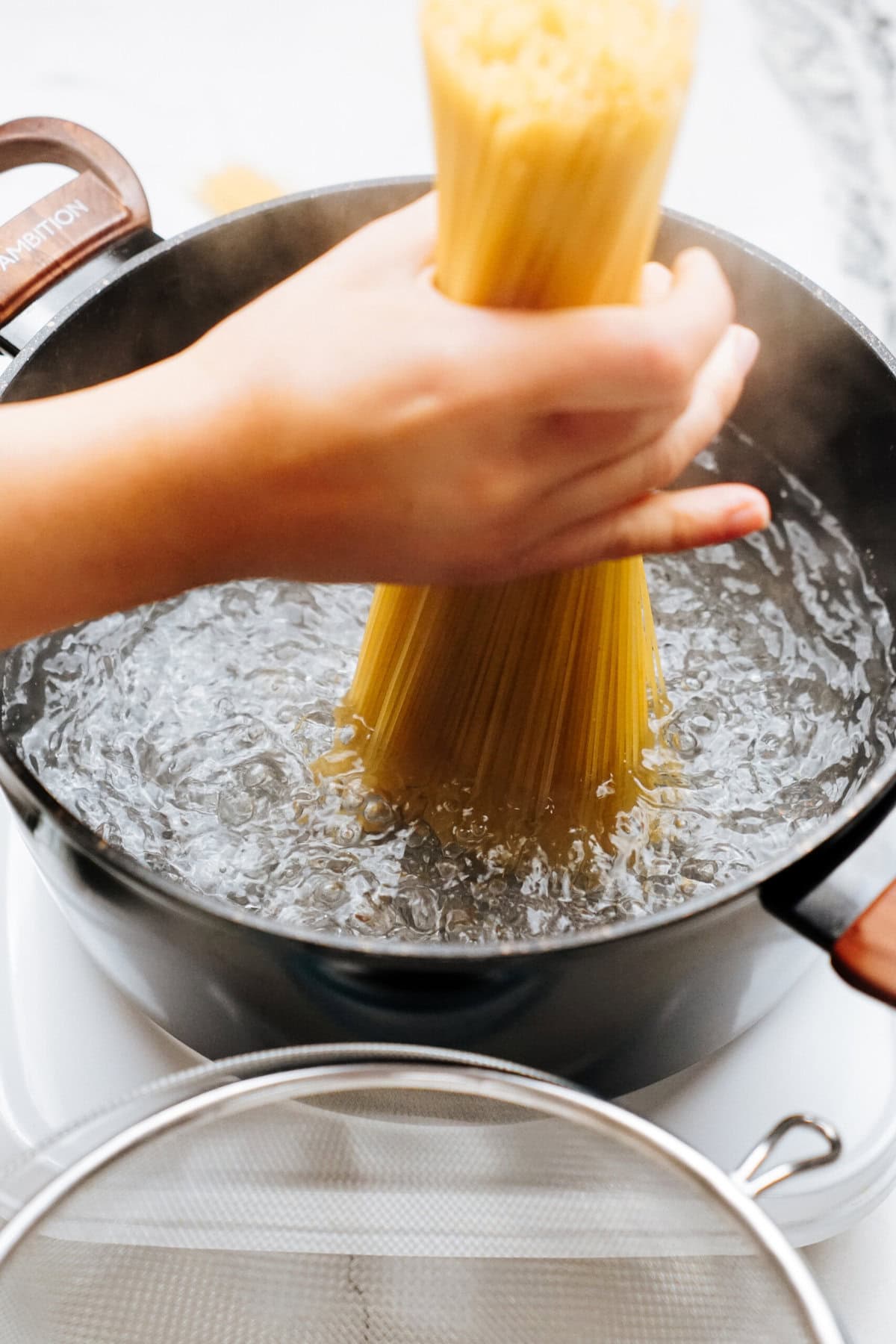 A hand is holding a bunch of uncooked spaghetti pasta above a pot of boiling water on a stove.