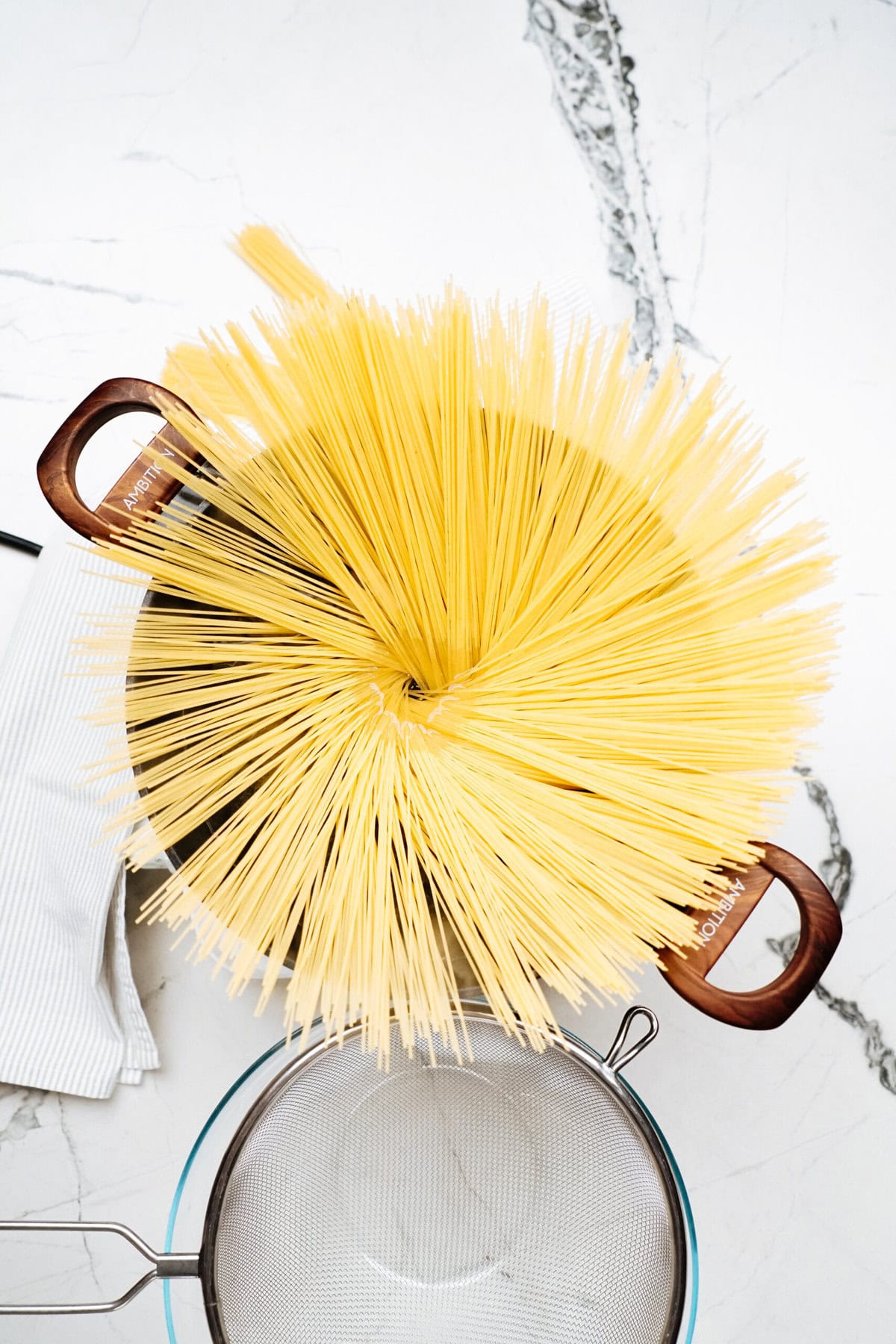 Overhead view of uncooked spaghetti arranged in a pot with handles, next to a mesh strainer on a marble countertop.