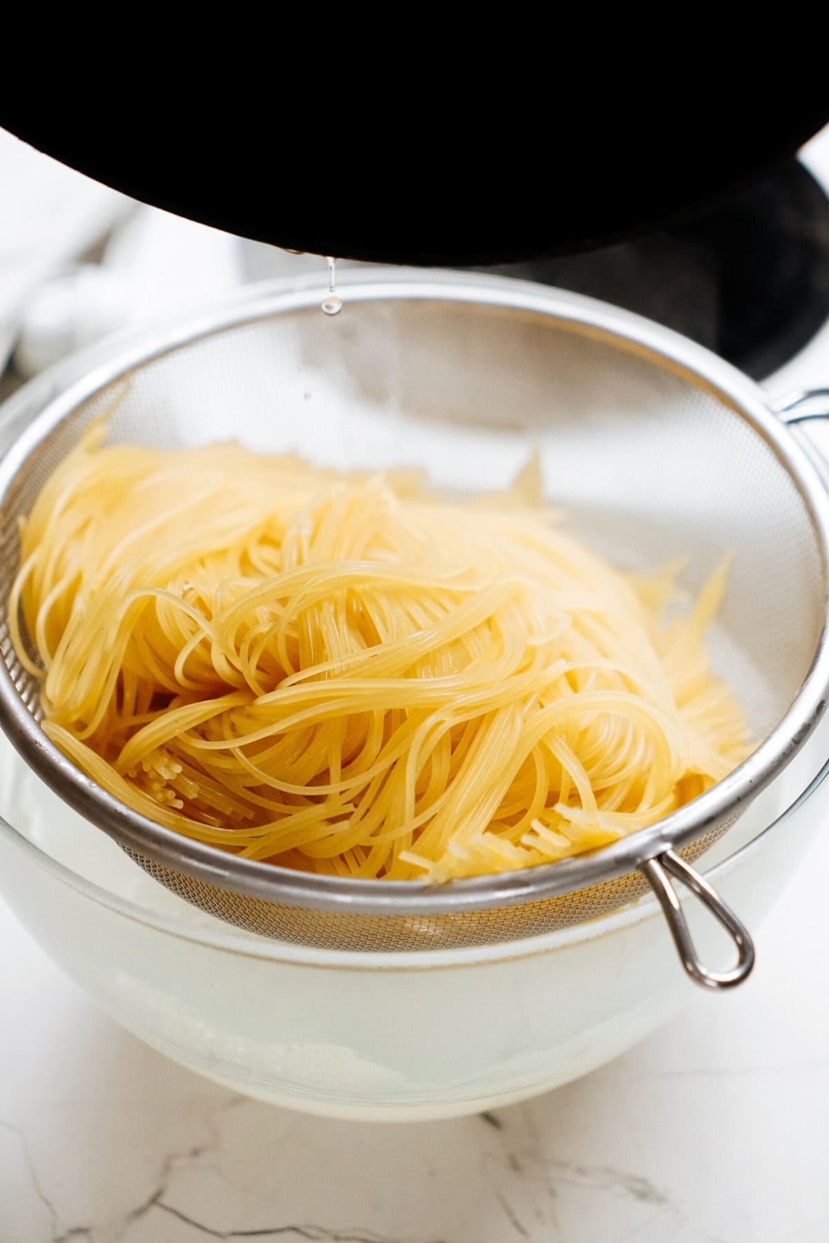 A close-up of cooked pasta being drained in a metal mesh strainer placed over a glass bowl.