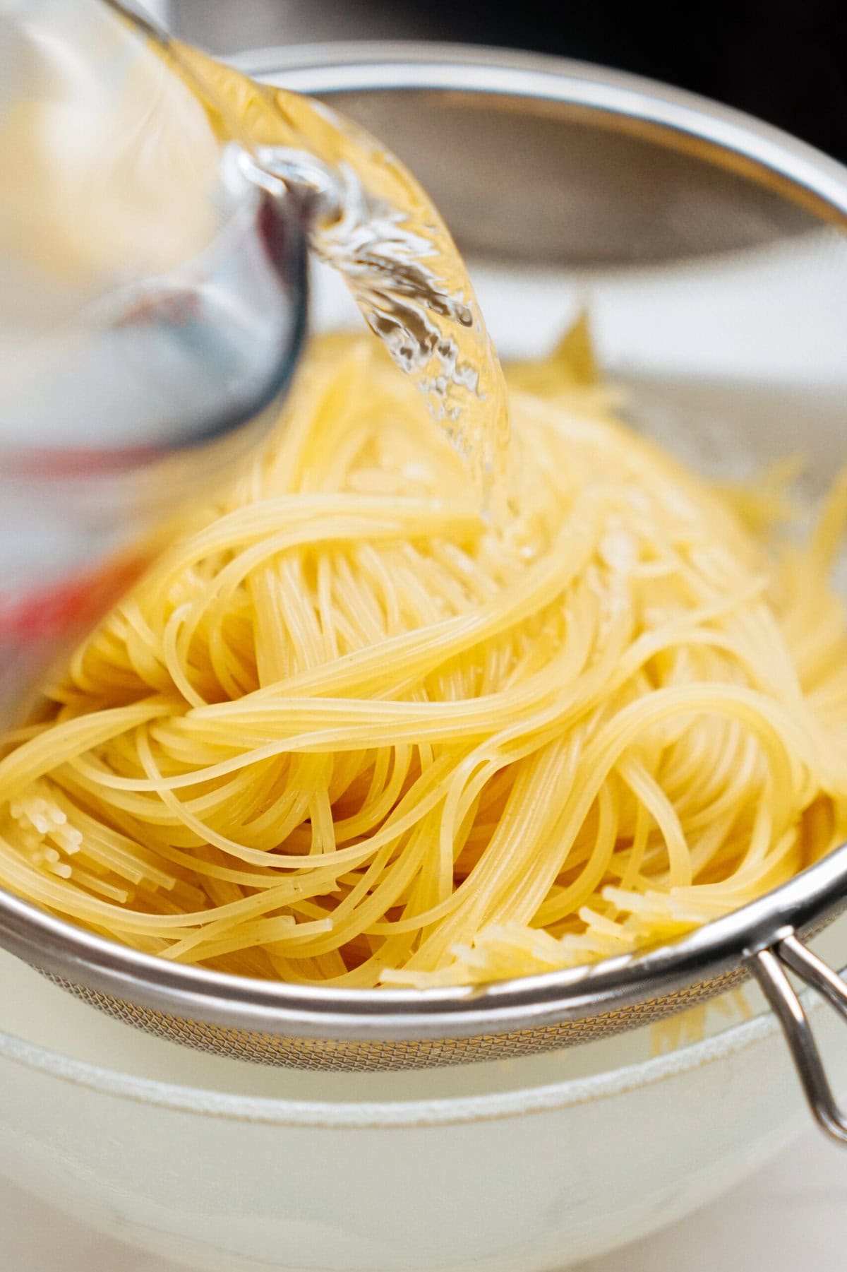 A metal strainer filled with cooked pasta is having water poured into it from a glass container.