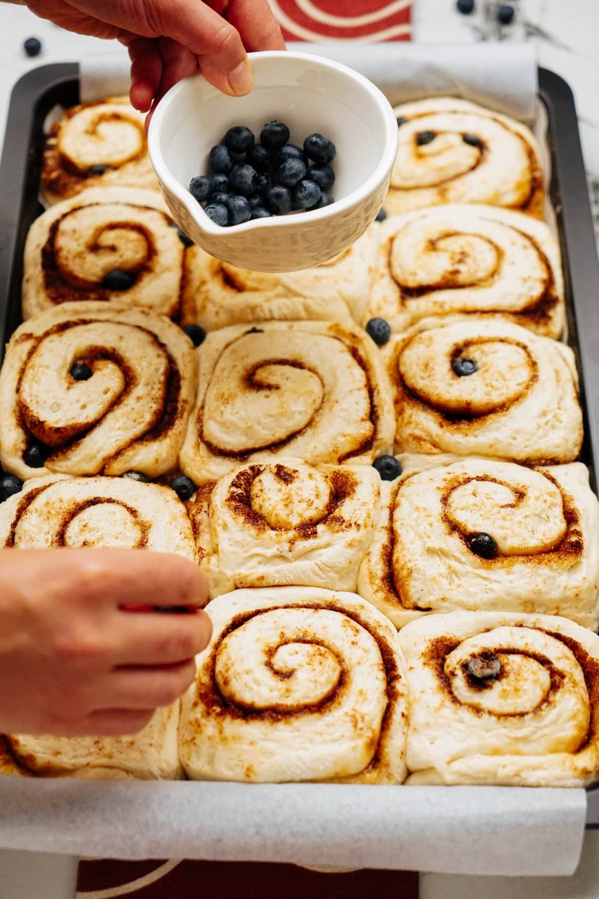 A person is placing blueberries into the center of a tray of unbaked cinnamon rolls, adding a delightful twist to the classic pastry.