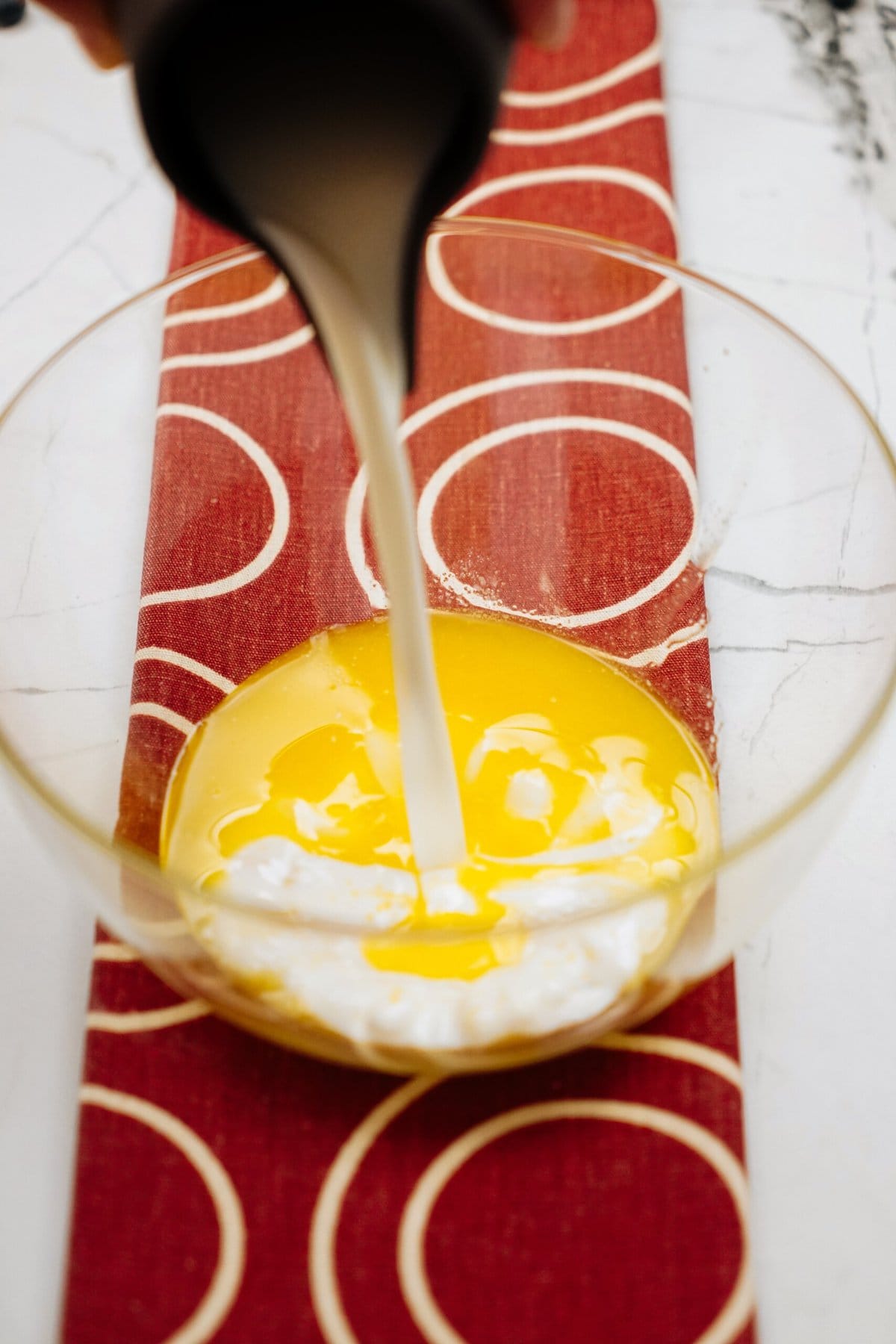 A liquid being poured into a glass mixing bowl containing beaten eggs on a red patterned surface, evoking the comforting aroma of freshly baked cinnamon rolls.