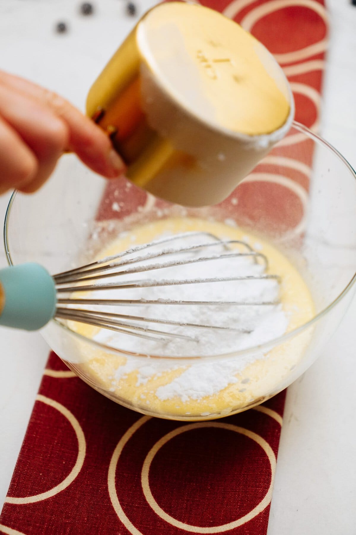 A hand is pouring powdered sugar from a measuring cup into a glass bowl containing a whisk and yellow batter, resting on a red and white patterned cloth, as if preparing for homemade cinnamon rolls.