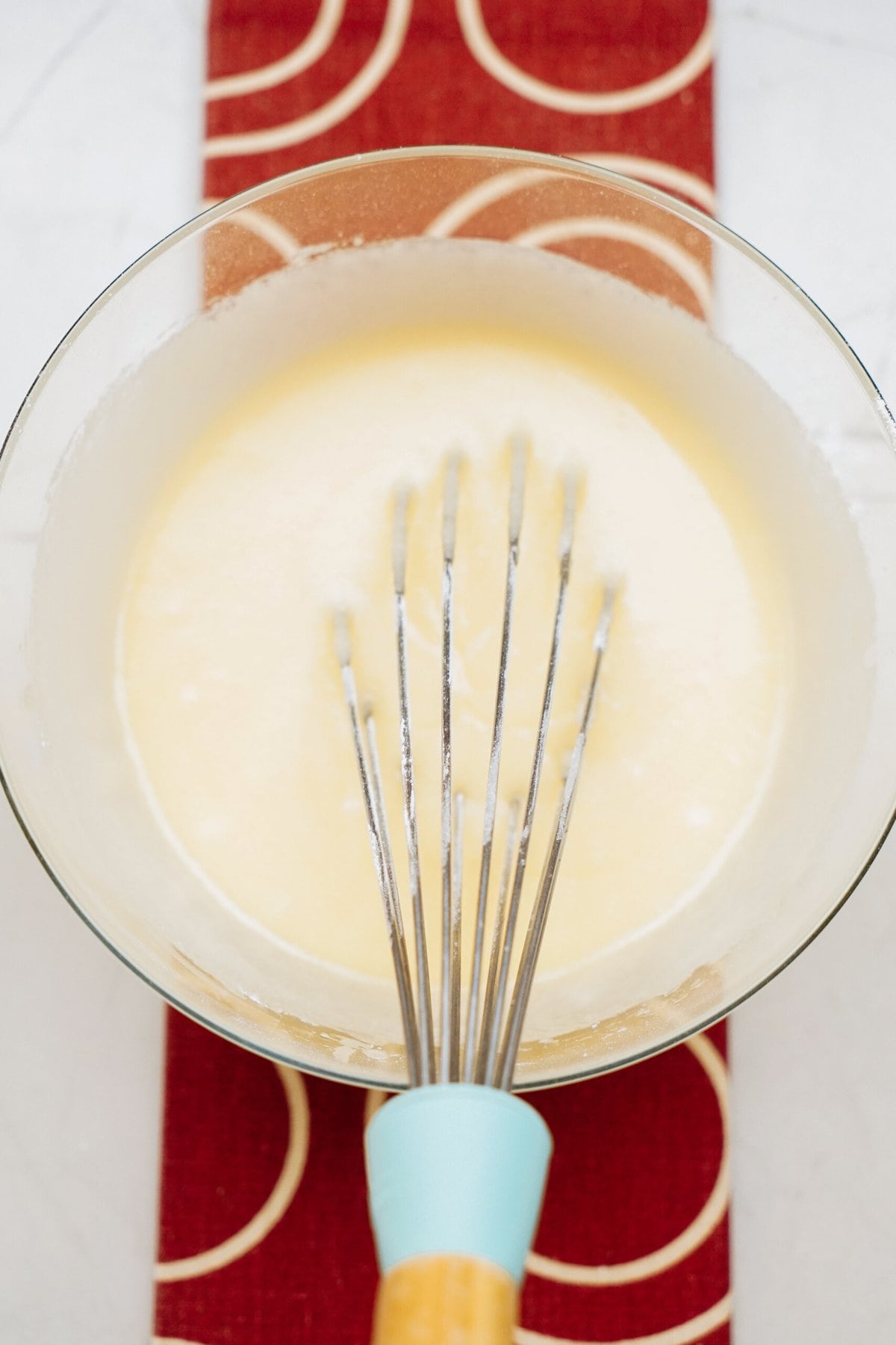 A whisk rests in a bowl of smooth, light yellow batter, reminiscent of the beginnings of delicious cinnamon rolls, placed on a red and white patterned surface.