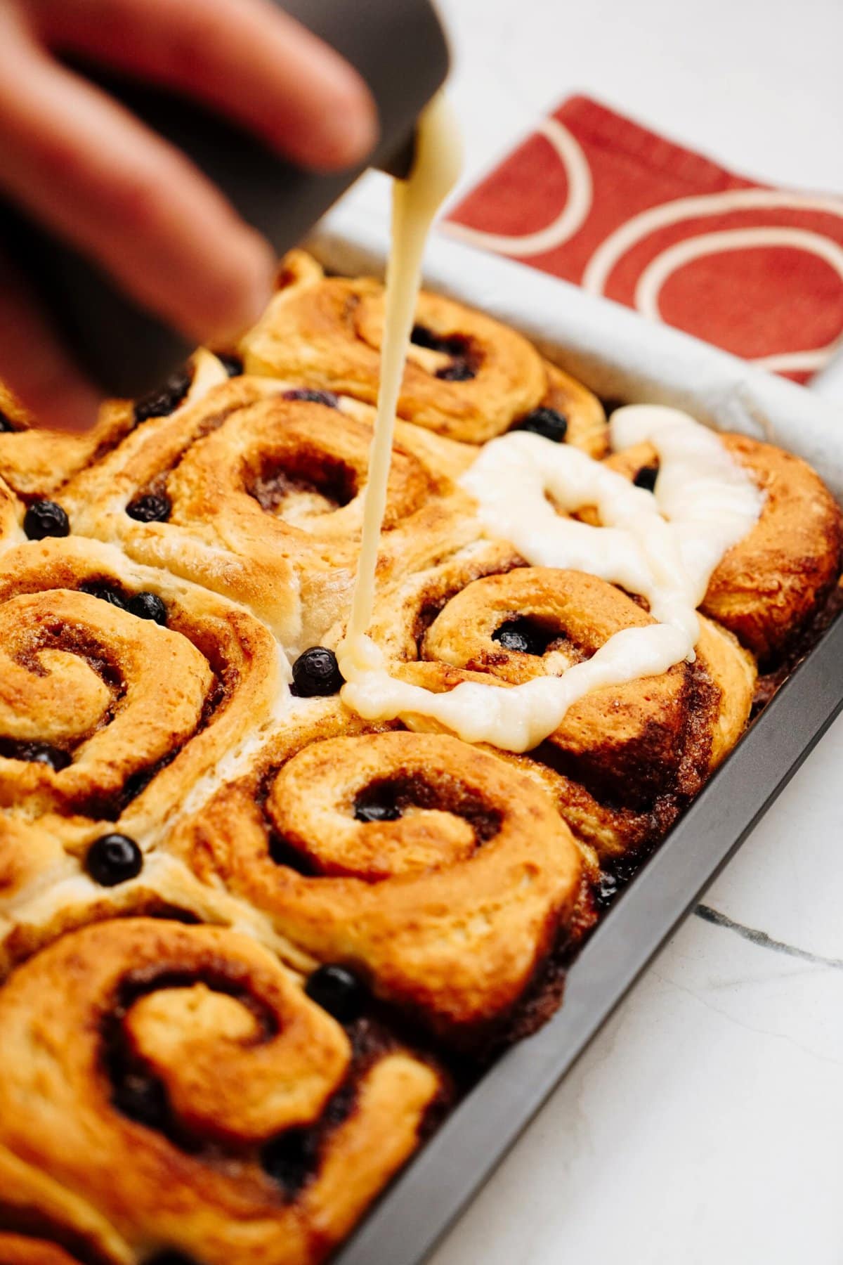 A hand is drizzling icing over a pan of freshly baked cinnamon rolls, filling the air with their sweet aroma.