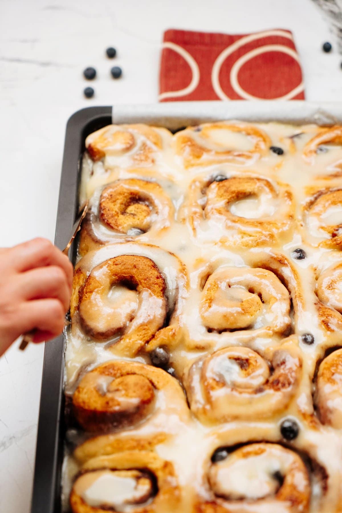 A person reaches to take a piece of glazed cinnamon roll from a baking tray with a red and white cloth in the background. Blueberries are sprinkled among the cinnamon rolls.