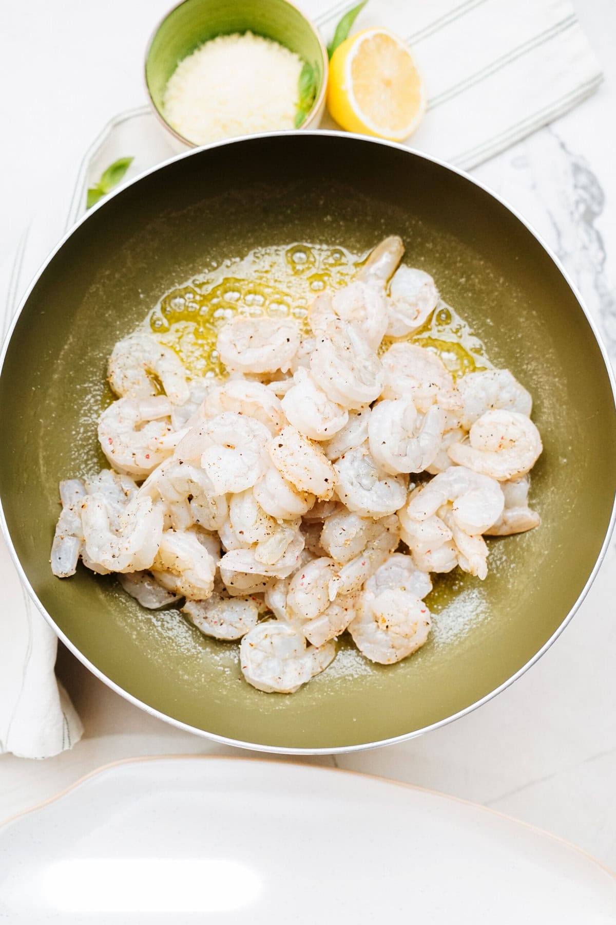 Shrimp seasoned with salt and pepper in a frying pan with oil, accompanied by a small bowl of grated Parmesan cheese, a lemon half, and a white cloth with green stripes.