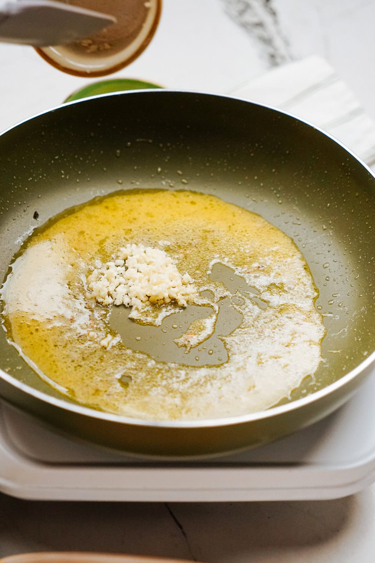 A frying pan with minced garlic sautéing in melted butter on a white stovetop.