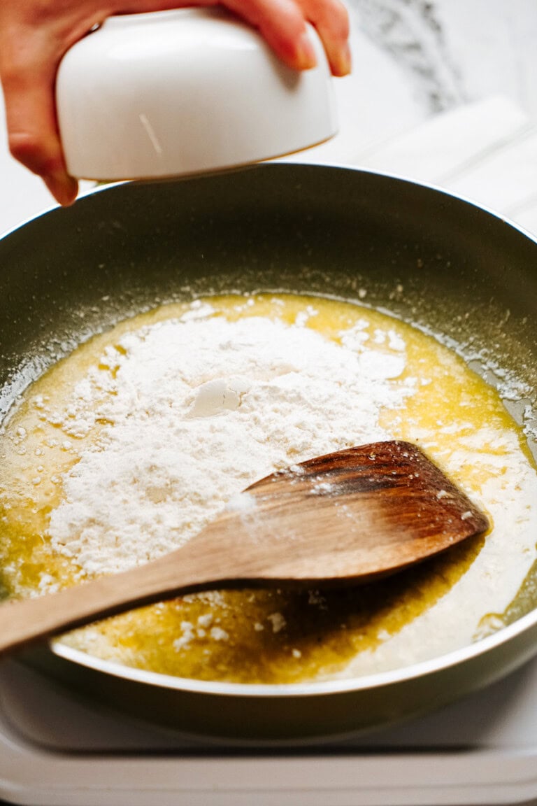 A person adds white flour from a small bowl into a pan with melted butter, while a wooden spatula rests in the mixture, indicating the initial steps of a cooking process.