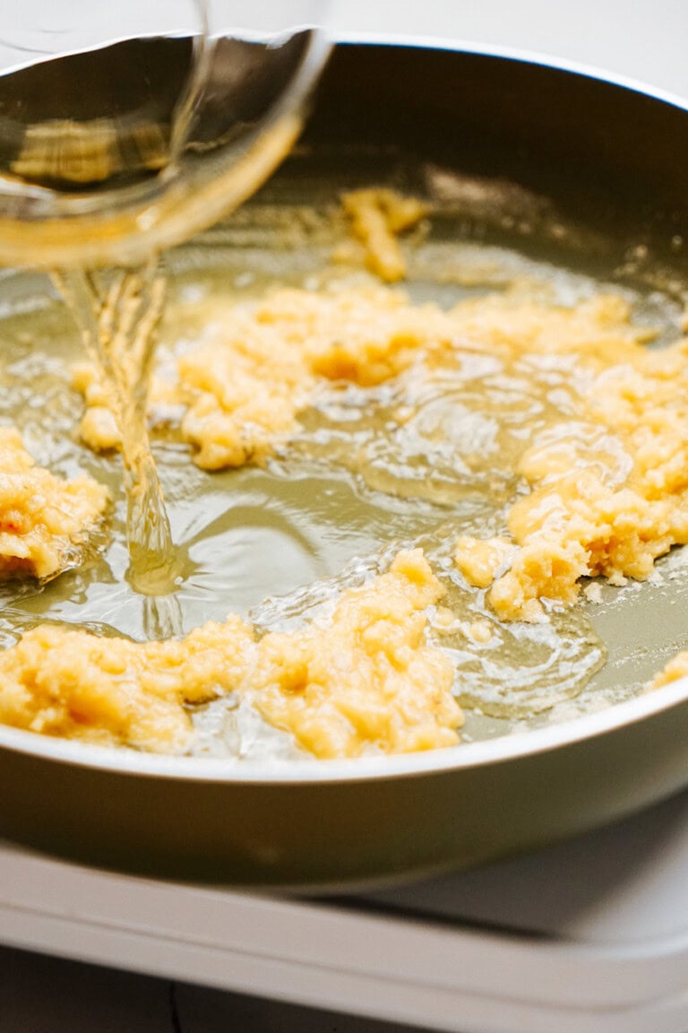 Water being poured from a glass bowl into a frying pan containing a partially cooked mixture.