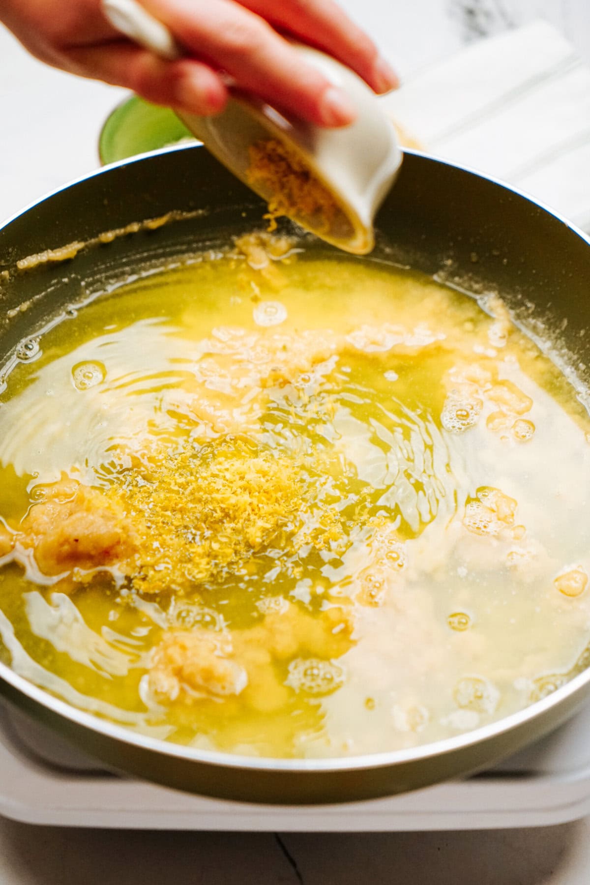 A hand is adding grated ginger to a frying pan filled with hot oil.