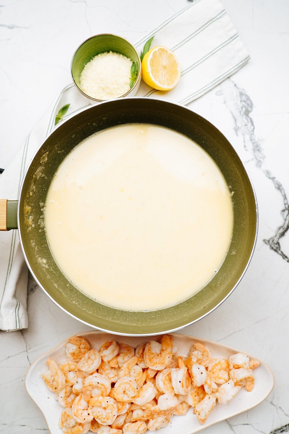 A pan of creamy sauce on a marble surface with a plate of cooked shrimp, a bowl of grated cheese, and a half lemon.