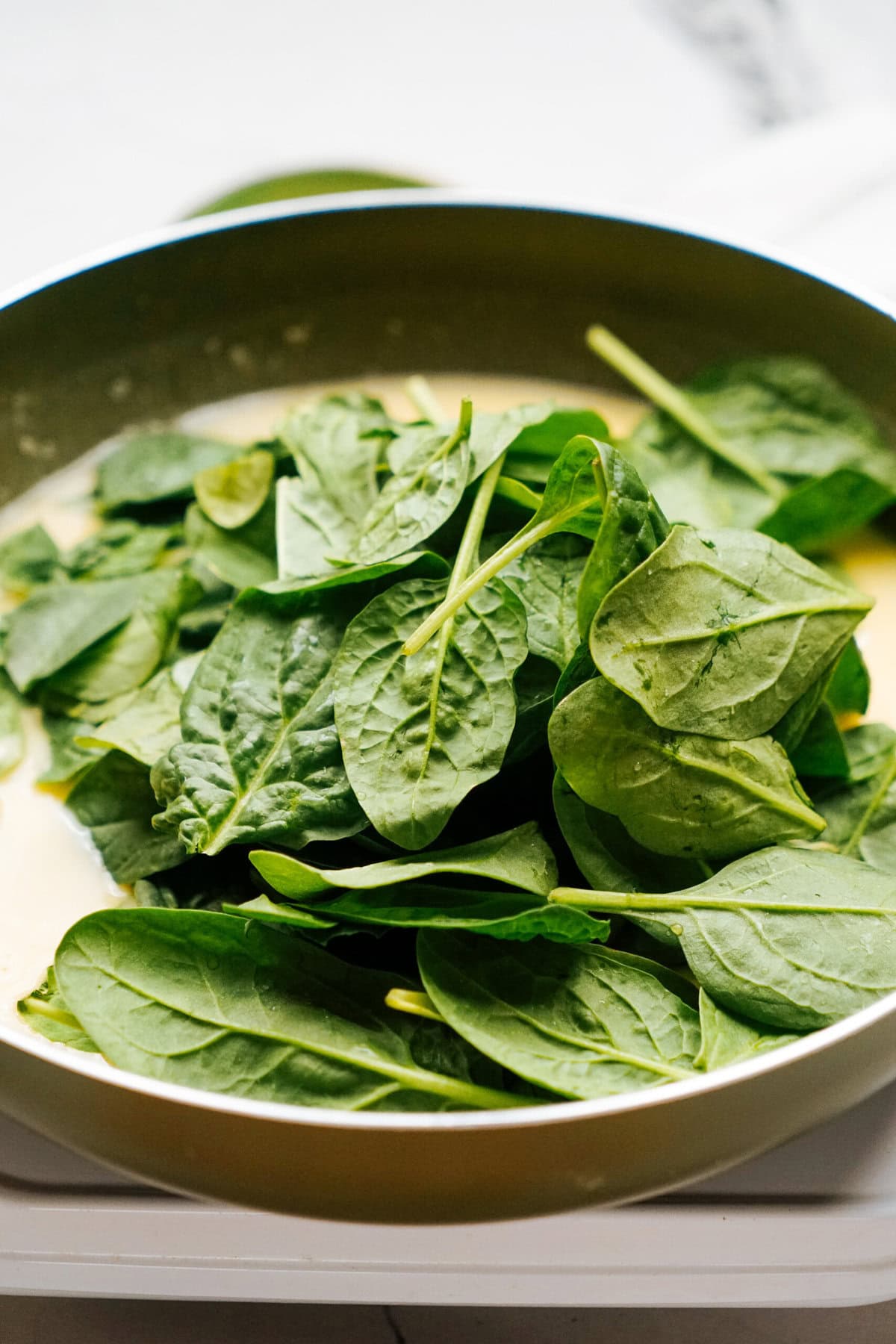 A frying pan filled with fresh spinach leaves on a white surface.