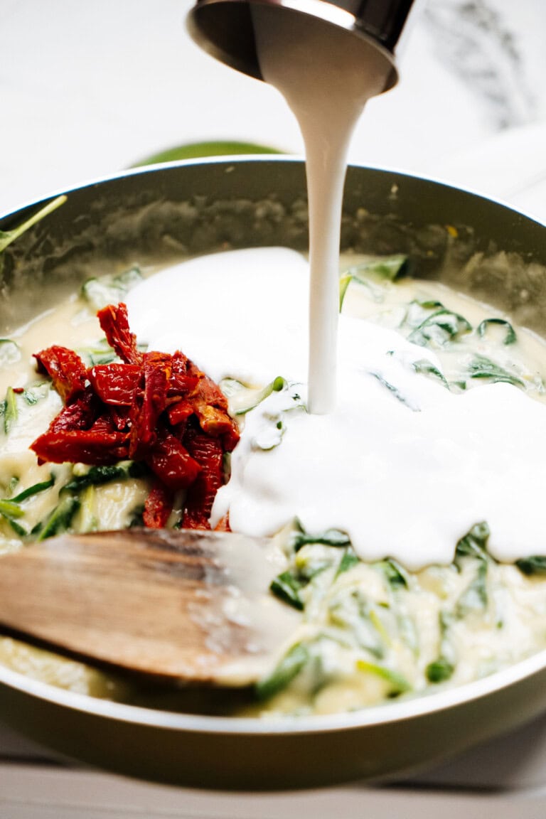 A green pan contains a creamy mixture with green vegetables and red peppers. Liquid is being poured from a container into the pan while a wooden spoon is partially submerged in the mixture.