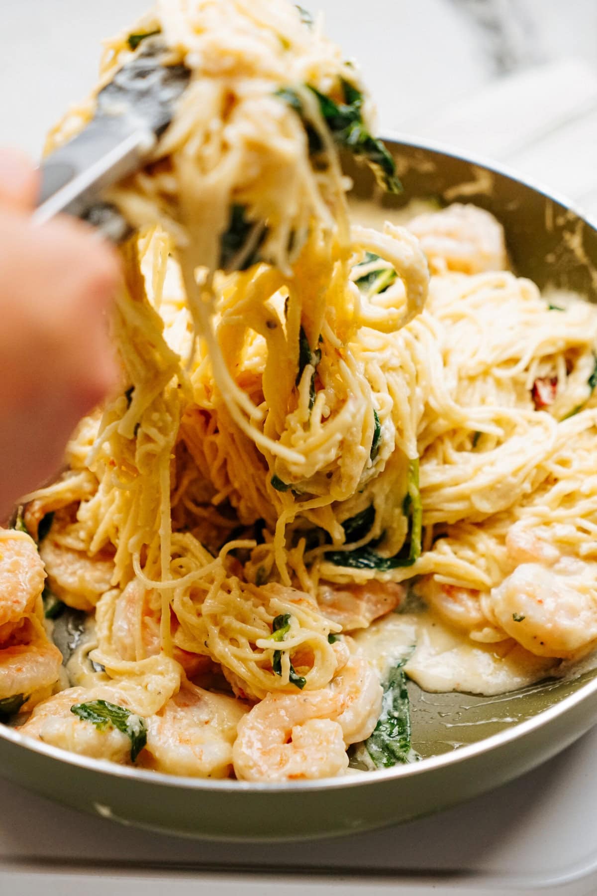 A person uses tongs to lift a portion of shrimp pasta with creamy sauce from a skillet.