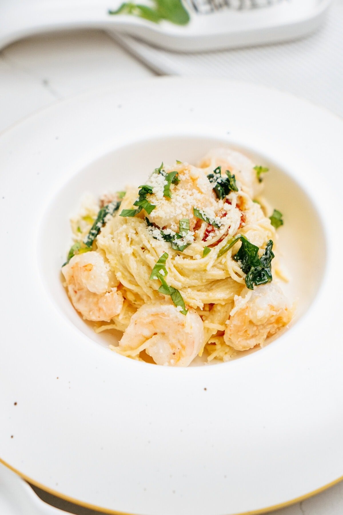 A plate of spaghetti with shrimp, parmesan cheese, and greens is served on a white dish.