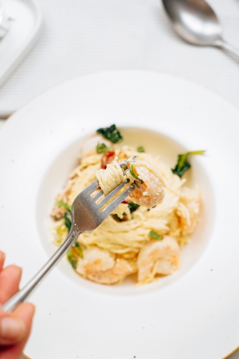 A hand holds a fork with a bite of pasta and shrimp above a white plate filled with more pasta, shrimp, and greens. A spoon is visible on the table in the background.