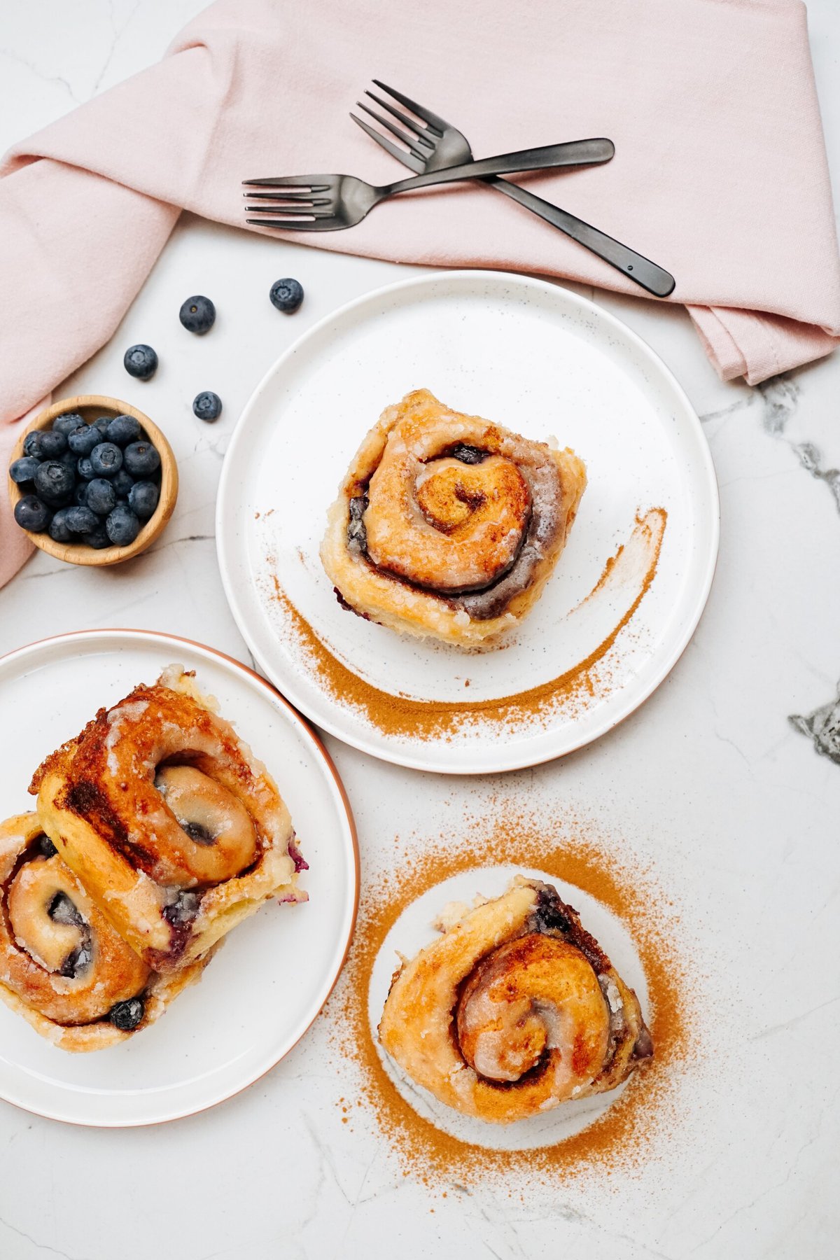 Top view of three cinnamon rolls on white plates, elegantly garnished with cinnamon powder. A small bowl of blueberries and two forks neatly placed on a pink napkin are positioned nearby.