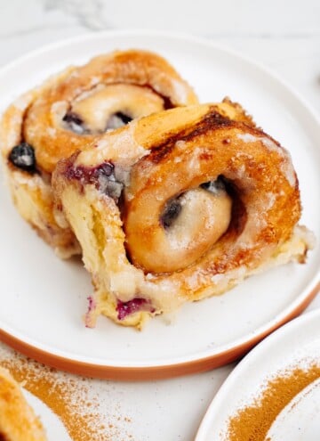 Close-up of two cinnamon rolls with a blueberry glaze on a white plate, with a light dusting of cinnamon on the plate's rim and a marble countertop in the background.