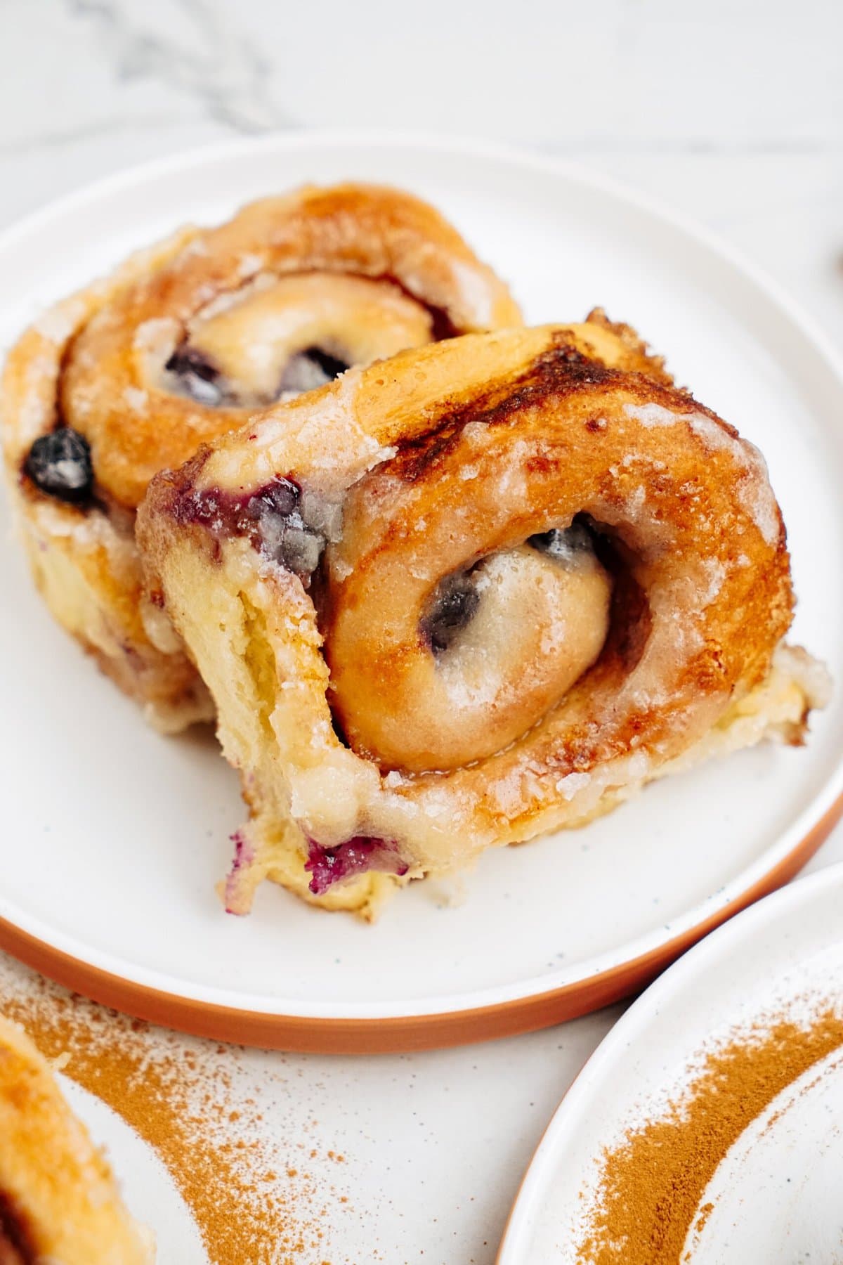 Close-up of two cinnamon rolls with a blueberry glaze on a white plate, with a light dusting of cinnamon on the plate's rim and a marble countertop in the background.