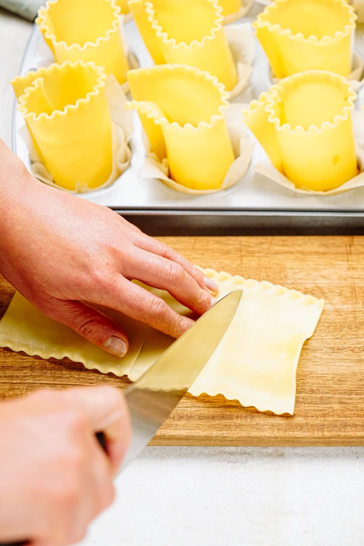 Hands cutting lasagna noodles on a wooden board with a knife. Rolled pasta sheets in muffin tin visible in the background, ready to shape into delicious lasagna cups.