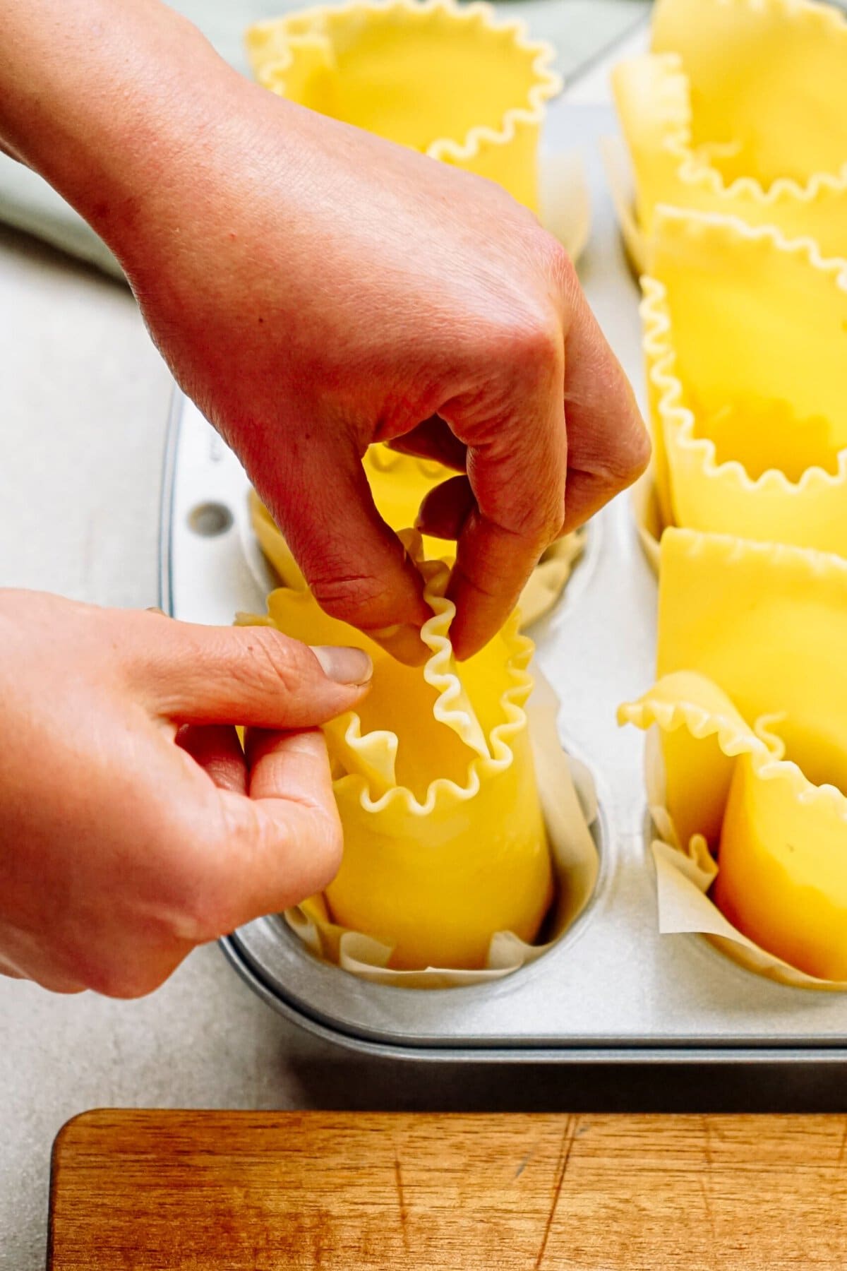 Hands assembling lasagna cups in a muffin tin, with uncooked lasagna noodles being shaped and placed individually.