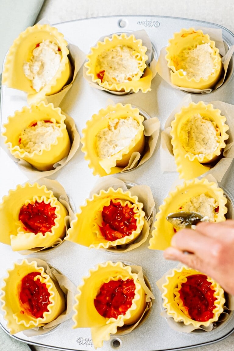 A metal muffin pan filled with vertical lasagna roll-ups, resembling bite-sized lasagna cups. Some are stuffed with a cheese mixture, while others are topped with red sauce. A hand holds a spoon near one of the rolls.