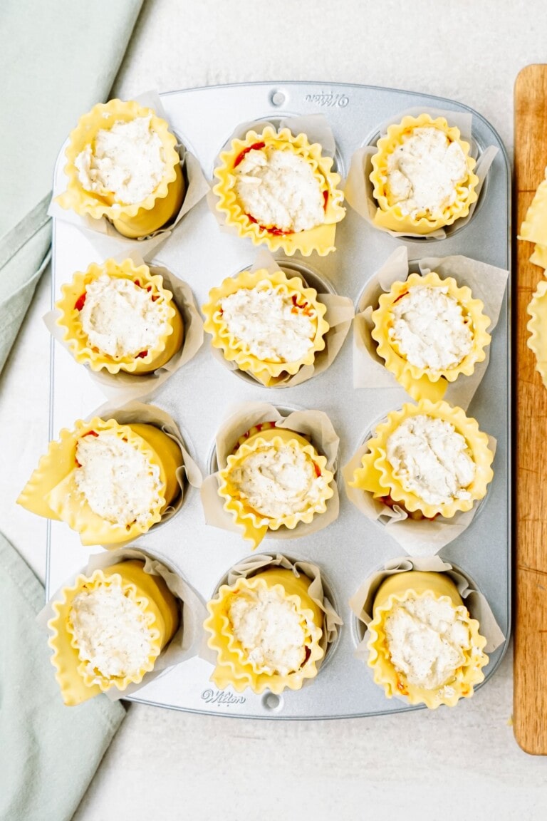 Twelve unbaked lasagna cups filled with cheese mixture are arranged in a muffin tin lined with parchment paper.