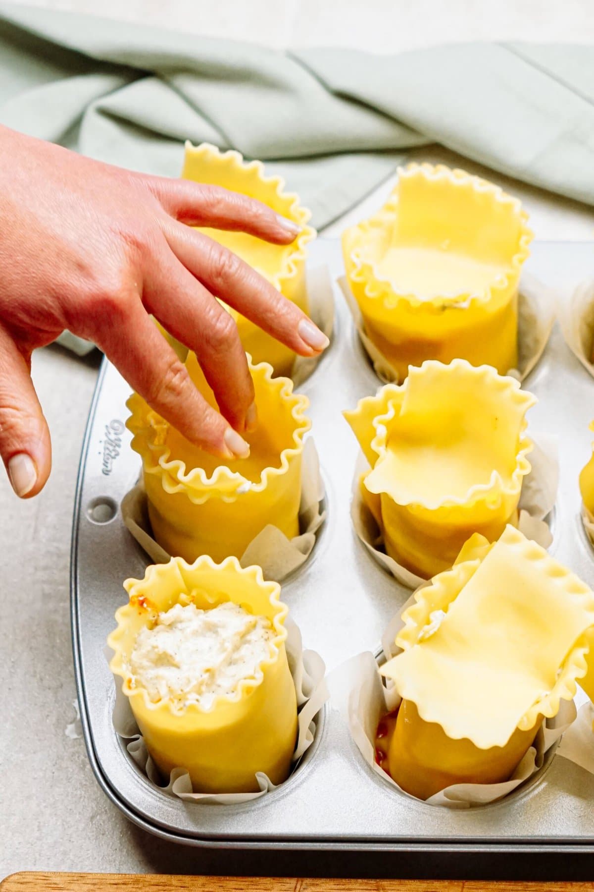A hand arranges lasagna roll-ups in a muffin tray, each lined with parchment paper and filled with cheese mixture, creating delightful lasagna cups.
