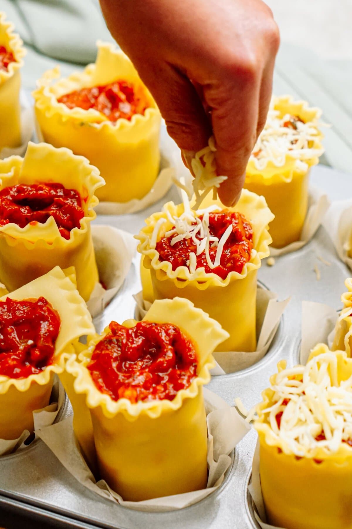 A hand topping individual lasagna roll-ups with shredded cheese in a muffin tin, creating delicious lasagna cups filled with rich tomato sauce.