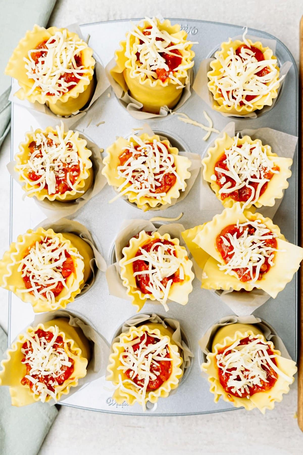 A muffin tin filled with lasagna cups topped with tomato sauce and shredded cheese, ready for baking.