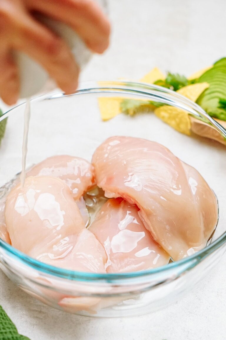 A hand pouring oil over raw chicken breasts in a clear glass bowl, with some green vegetables and yellow objects in the background.