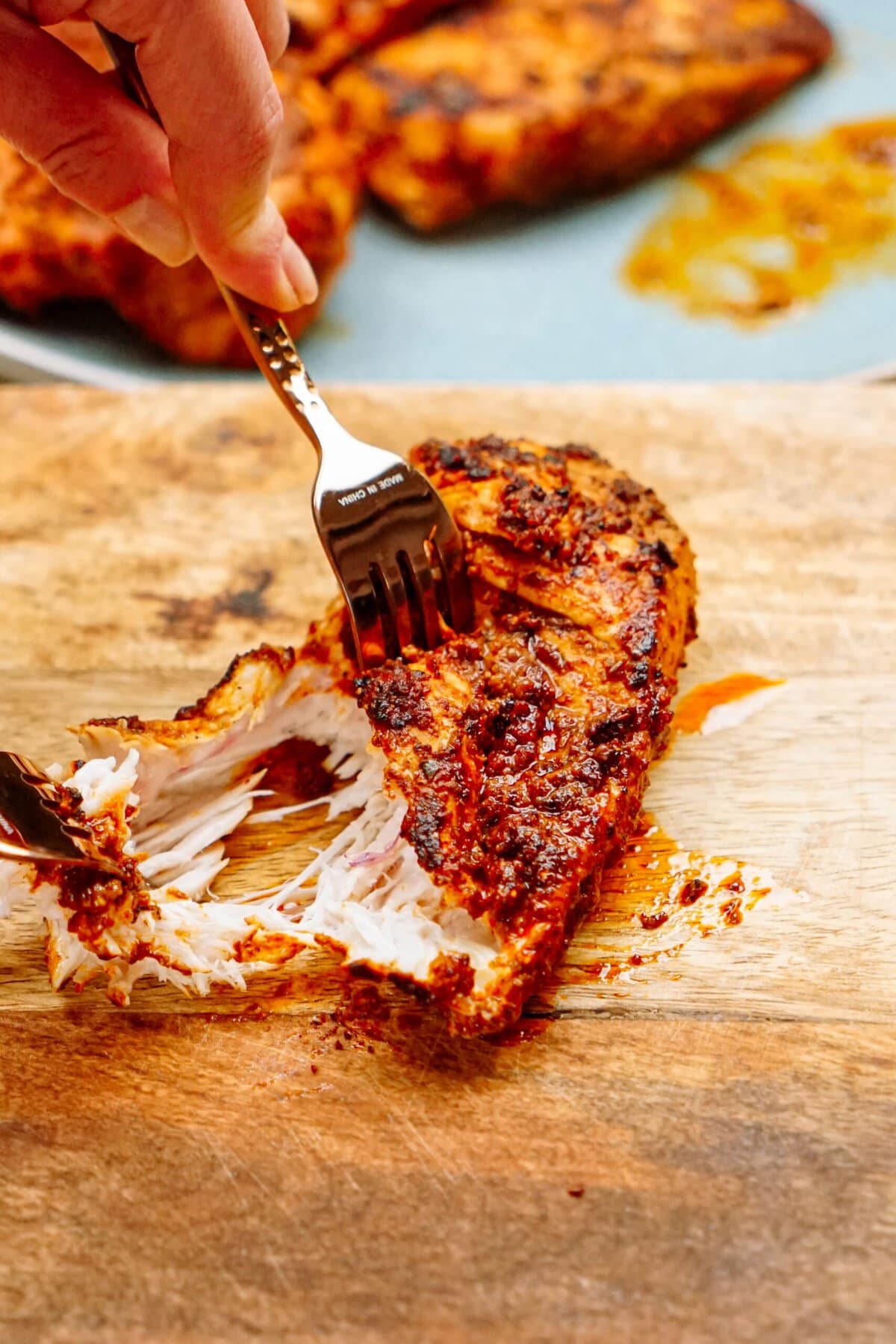 Close-up of a fork pulling apart a piece of seasoned, grilled chicken breast on a wooden cutting board. Another piece of meat is blurred in the background.