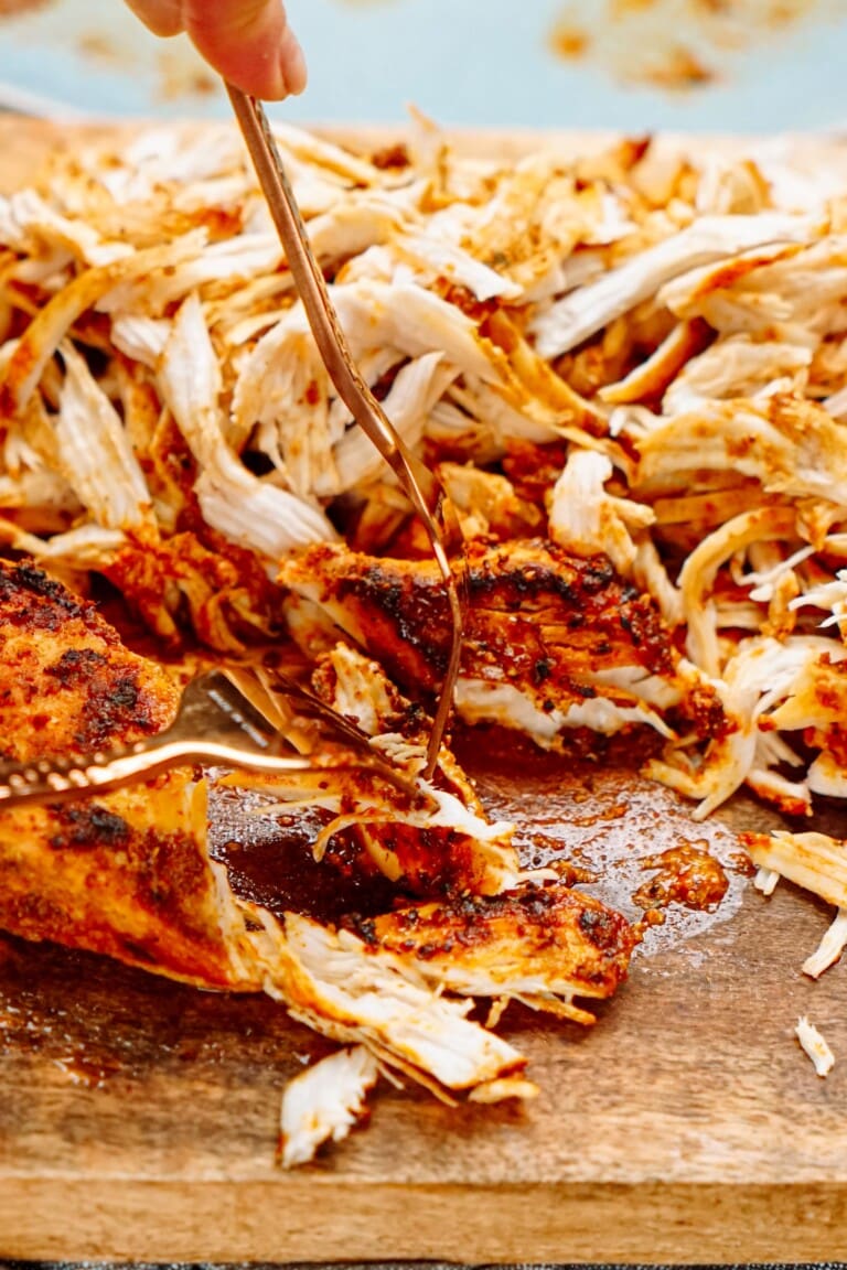 A fork shredding seasoned, cooked chicken on a wooden cutting board.