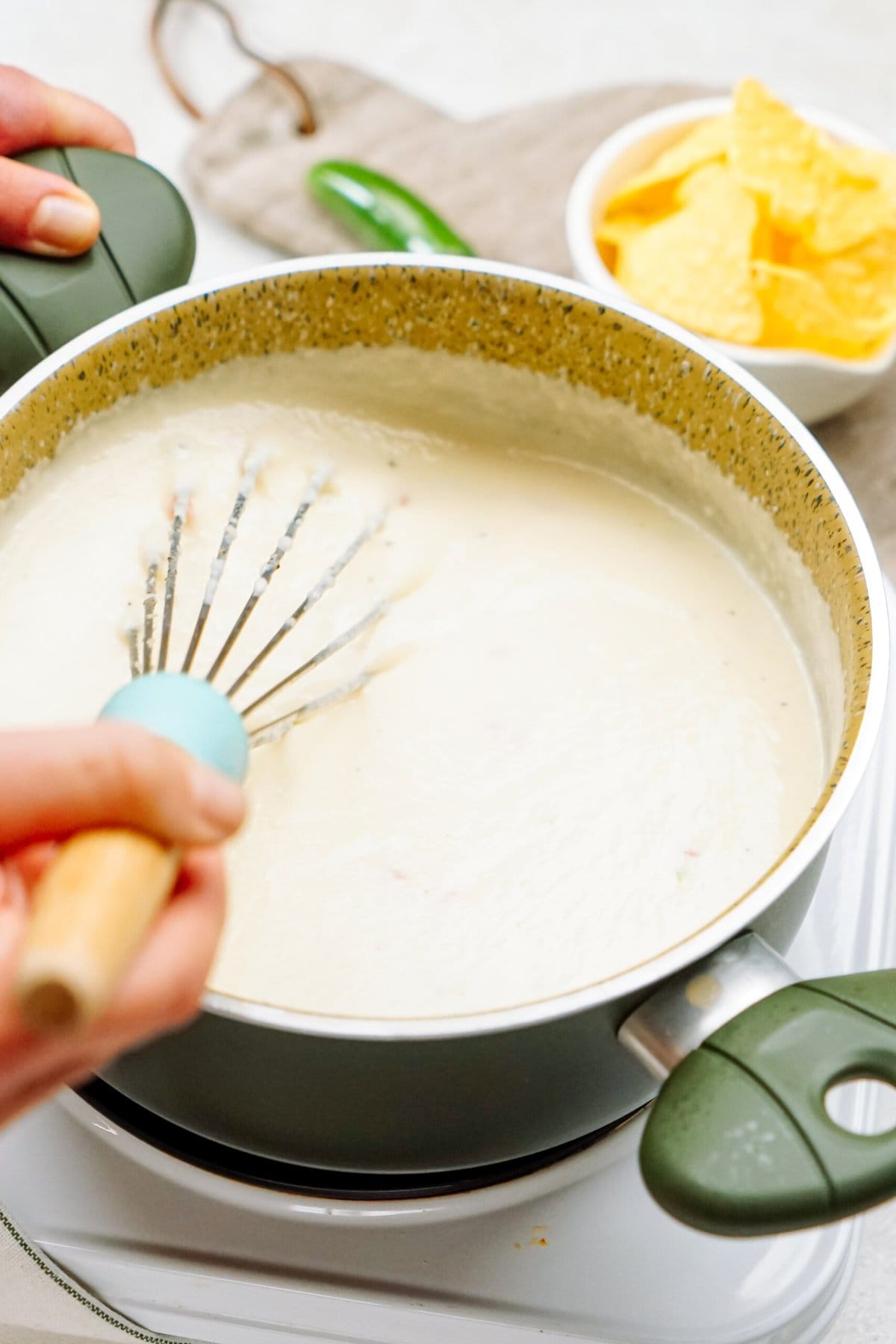 Hands are shown stirring a creamy white queso with a whisk in a green saucepan on a stove. A bowl of tortilla chips is in the background.