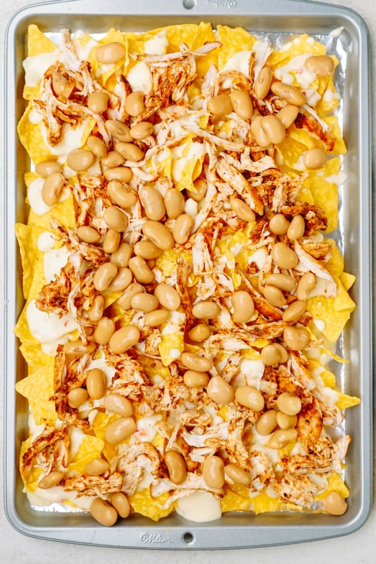 A baking tray filled with tortilla chips topped with shredded chicken, white beans, and melted cheese.