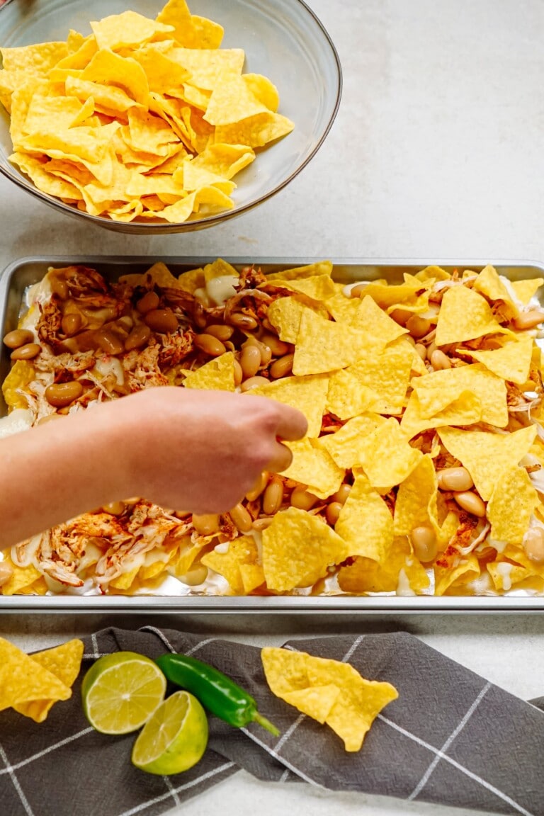 A person assembling nachos on a baking sheet with chips, beans, and chicken. A bowl of chips, a sliced lime, and a green pepper are on the counter nearby.