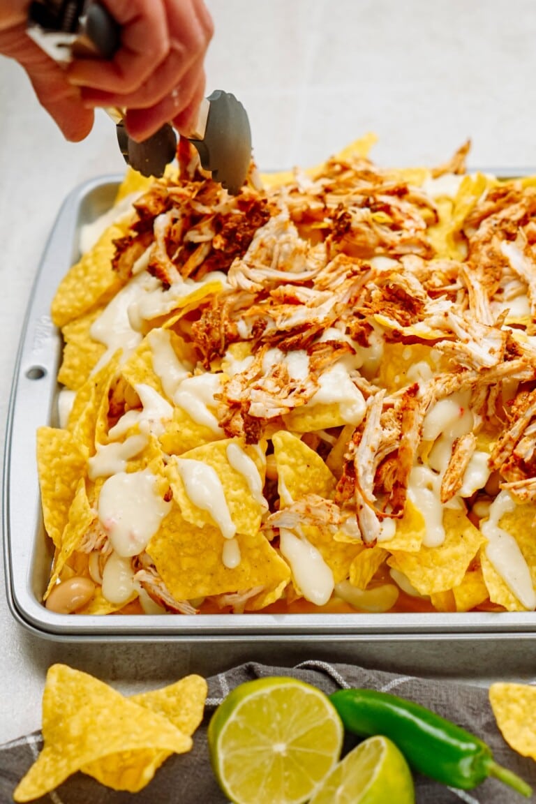 A tray of nachos topped with shredded chicken and cheese sauce. A hand with tongs is arranging the toppings. Sliced lime and whole jalapeños are visible in the foreground.