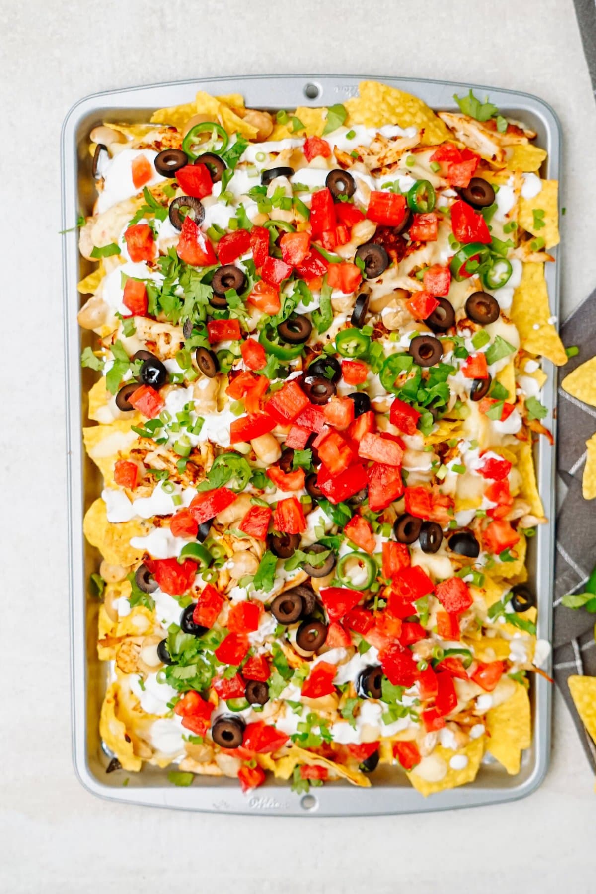 A tray of loaded nachos topped with melted cheese, diced tomatoes, black olives, jalapeño slices, chopped cilantro, and drizzled with sour cream. Corn tortilla chips are visible at the edges of the tray.